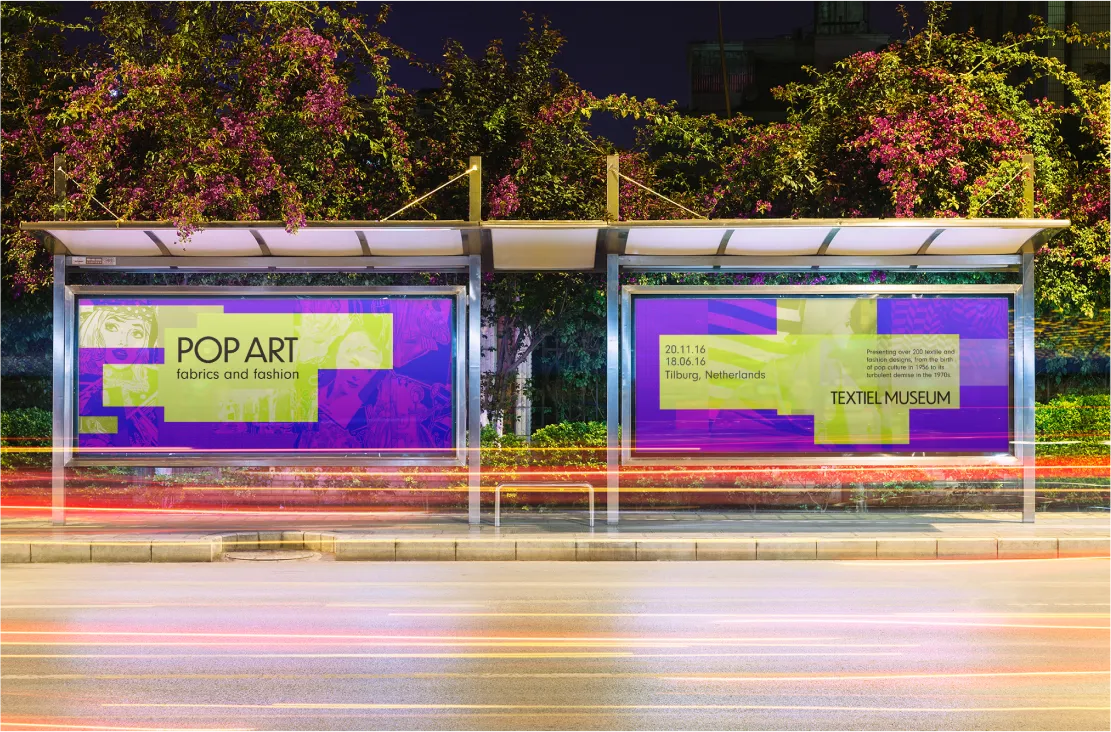 Photo-realistic mockup of a lit bus-stop shelter at night, with two 'Pop Art fabrics and fashion' Textiel Museum ad panels, long-exposure light trails from passing traffic.